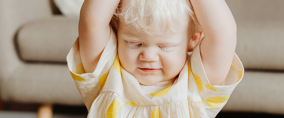 Blondes Kleinkind, weiblich, in weißem Kleid mit großen gelben Punkten, das sich mit erhobenen Armen ein Taschentuch über den Kopf hält. Auf ihrem Kleid und neben ihr liegen Spielzeugelemente aus Kunststoff. Im Hintergrund ist ein beiges Sofa zu sehen.